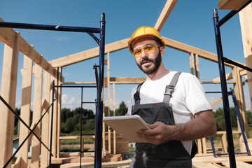 The man is a builder on the background of the roof of a frame house, in a yellow helmet and gray...