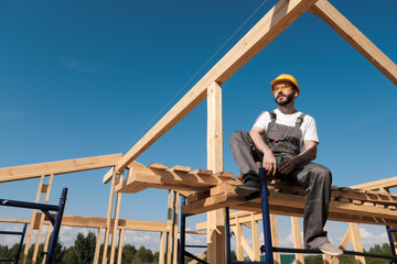 The man builder sits on the edge of the roof of the frame house, in a yellow helmet and gray...