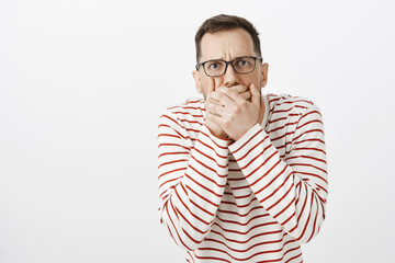 Fototapeta premium Guy want throw up tasting awful food. Portrait of displeased european boyfriend in black glasses, covering mouth with both palms and frowning, suffering from stomachache and wanting vomit
