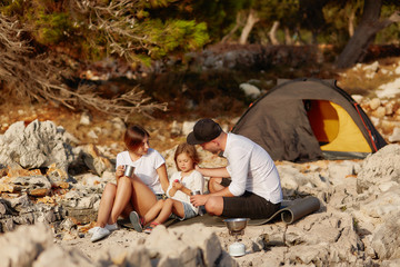 Friendly family, sitting near tent on stone seacoast at daytime.