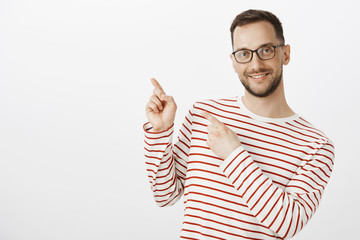 Naklejka premium Cute tender adult male gay model in glasses, pointing at upper left corner with finger gun gestures and smiling with pleased friendly expression, showing lgbt place to friend over gray wall