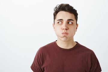 Fototapeta premium Portrait of funny european male model in red t-shirt, holding breat, sulking and looking left while holding water in mouth, standing over gray background during appointment in dentist office