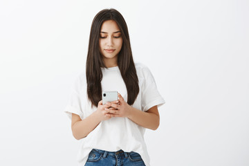 Writing important message to girlfriend. Calm good-looking european woman in casual outfit, holding smartphone and typing while looking at screen with slight happy smile, standing over gray wall