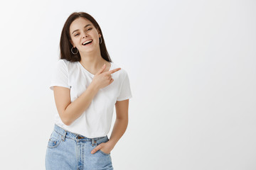 Charming hot and stylish dark-haired female in stylish street style outfit, holding hand in pocket and pointing right with forefinger, laughing joyfully, standing happily over gray background