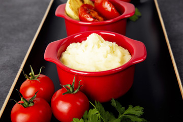 Food is still life. Two red pots with potato puree and fried vegetables on a black tray. Background black marble.