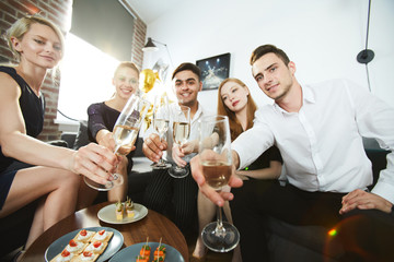Circle of young people toasting with flutes of champagne over served table with starters and appetizers