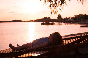 Small boy relaxing on deck chair by the water at sunset.