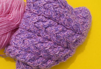 Coats of wool pink and purple lie next to an unconnected cap. Yellow background.
