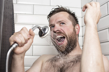 Cute bearded man singing in the bathroom using the shower head with flowing water instead of a...