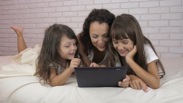 Family in the bedroom with a tablet. Mom with daughters on the bed with a tablet.