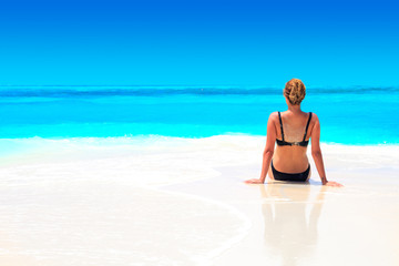 Blond woman with black bikini tans on the sandbank in front of turquoise ocean and blue sky