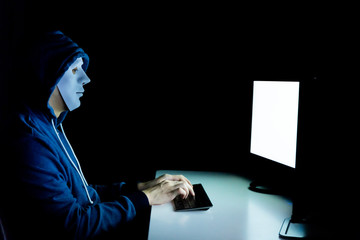 Masked hacker under hood using computer to hack into system and trying to commit computer crime -...