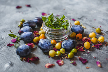 a jar of mint and scattered blue and yellow plums on a gray background