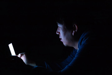 Asian young man using phone in dark bedroom with blue light phone screen - unhealthy eyes strain concept.