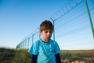 upset poor little boy refugee with dirty face near wall fence on border