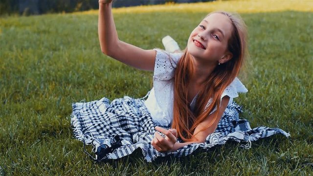 Young beautiful girl making selfie portrait using smartphone in green park, lying on the grass with mobile phone. Color graded
