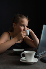 A teenager girl in glasses works at a computer at night and drinks coffee on a dark background with copy space, a concept of freelancing