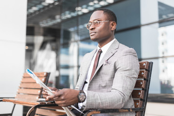 black man businessman in a business suit and glasses sits on a bench and reads a newspaper against the backdrop of a modern city