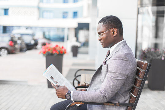 black man businessman in a business suit and glasses sits on a bench and reads a newspaper against the backdrop of a modern city