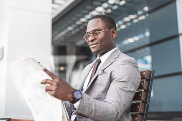 Fototapeta premium black man businessman in a business suit and glasses sits on a bench and reads a newspaper against the backdrop of a modern city
