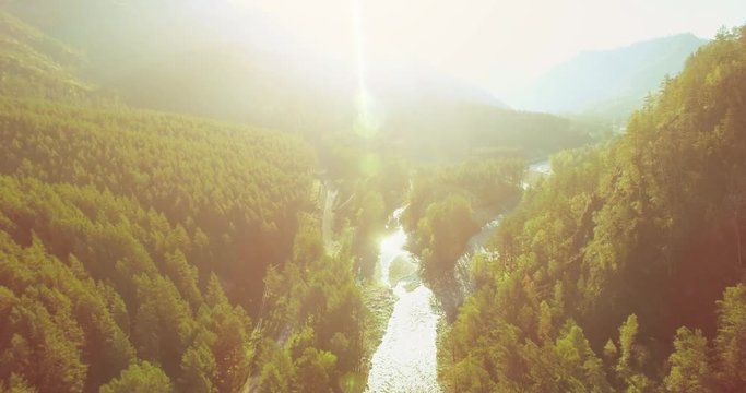 Aerial UHD 4K view. Mid-air flight over fresh mountain river and meadow at sunny summer morning. Green trees and sun rays on horizon. Rural dirt road below.