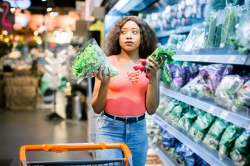 Pretty Afro young woman in jeans shopping organic veggies and fruits. Woman with trolley holding lettuce and radish in her arms