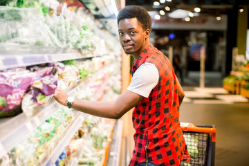 African man shopping in vegetable section at supermarket. Black man doing shopping at market while buying vegetables. Handsome guy holding shopping basket reading nutritional values of product.