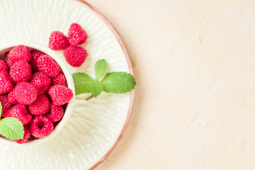 Ripe raspberries with green mint leaves in cup and saucer on pastel yellow background with copy space.