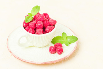 Ripe raspberries with green mint leaves in cup and saucer on pastel yellow background.