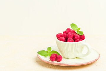 Ripe raspberries with green mint leaves in cup and saucer on pastel yellow background with copy space.