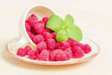 Ripe raspberries with green mint leaves in cup and saucer on pastel yellow background.