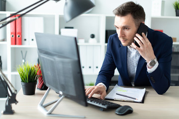 Obraz premium Młody mężczyzna stoi przy stole w biurze, rozmawia przez telefon i wpisuje tekst na klawiaturze.