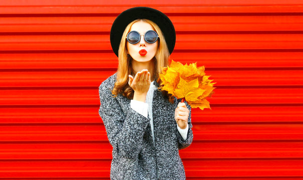 Stylish funny autumn woman sends an air kiss with yellow maple leaves, wearing a coat on red background