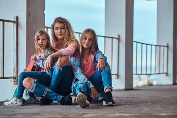 Beautiful girl with blonde hair and her little sisters dressed in trendy clothes sitting together on skateboards posing near a guardrail against a sea coast.