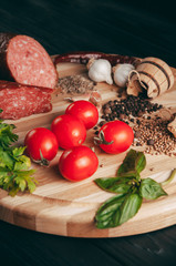 Smoked salami on a black table and a brown board. Sausages with rosemary, garlic and pepper and tomato cherry, dill, parsley.