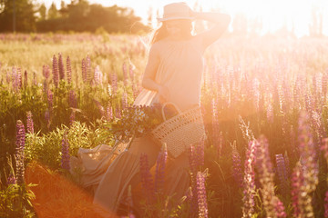 Woman holding wicker bag in her hands wearing fedora hat on sunset in lupine field