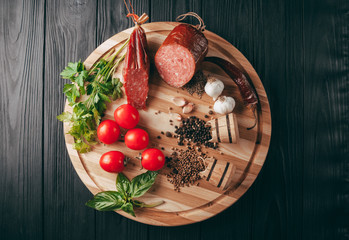Smoked salami on a black table and a brown board. Sausages with rosemary, garlic and pepper and tomato cherry, dill, parsley.