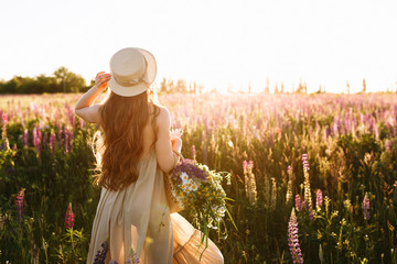 Young woman in straw hat and dress with bouquet of lupine flowers, back view in sunset field