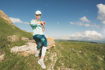 Fototapeta premium Portrait of a smiling young fitness girl in a cap and headphones checking her smart clock sitting on a rock outdoors against a blue sky with clouds
