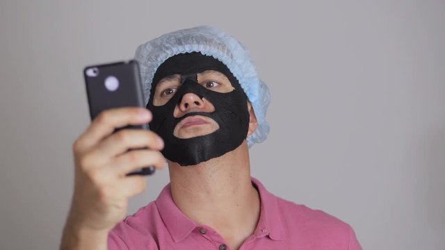 A man, applying a rejuvenating cosmetic mask of black cloth to his face and doing selfie with a smartphone