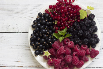 Red and black currants and raspberries and mint, useful ripe berries on a plate, wooden white background