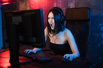 Plakat Screaming emotional young angry woman playing on personal computer holding game keyboard and mouse sitting on a chair at home. Gaming gamers concept.