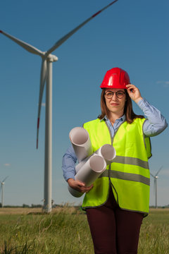 Portrait of a serious cute young female engineer in a green waistcoat and red hard hat with geodesic plans in hand against the backdrop of a windmill and blue sky.