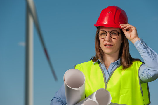 Portrait of a beautiful young female engineer in red helmet and glasses with projects and plans against the background of a windmill and blue sky