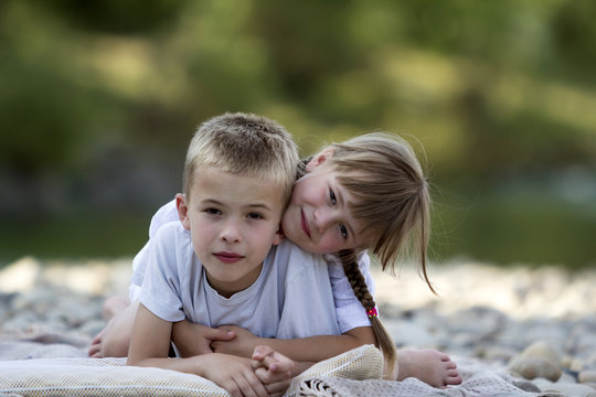 Two young happy cute blond smiling children, boy and girl, brother and sister laying embraced on pebbled beach on blurred bright sunny summer day background. Friendship and perfect holiday concept.