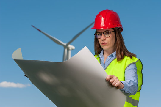 Serious young female engineer in red helmet and glasses with projects and plans against the background of a windmill and blue sky