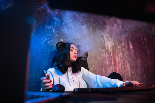 Young brunette woman wearing hoodie, sitting down by desk with computer while smoking e-cigarette