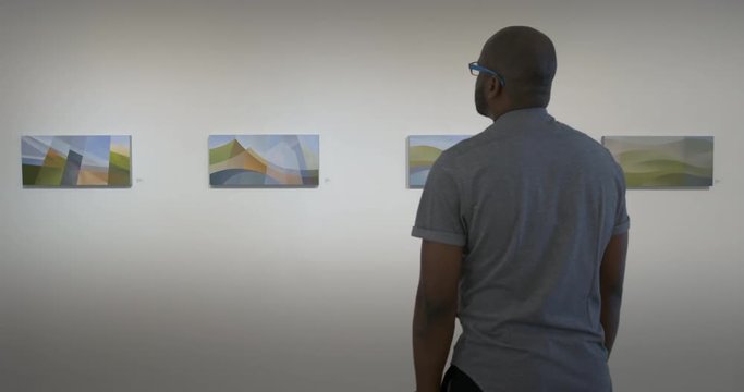 MS African American man walks into view and looks at four abstract paintings in art gallery. Locked off shot behind subject