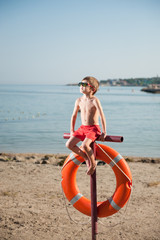 Naklejka na ściany i meble little boy in sunglasses sits on pole with orange lifebuoy hanging on beach