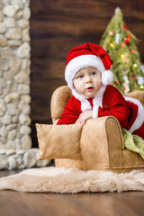 Close-up portrait of a Little boy dressed as a red Santa Claus, playing by a stone fireplace with bright garlands, putting a gift box under the Christmas tree. New Year card. The baby laughs, has fun.